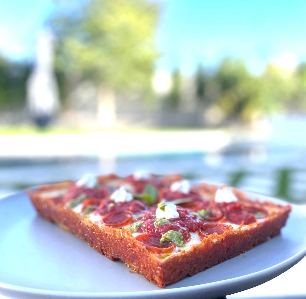 A slice of Detroit-style pepperoni pizza on a blue plate, with a backyard pool blurred behind it.