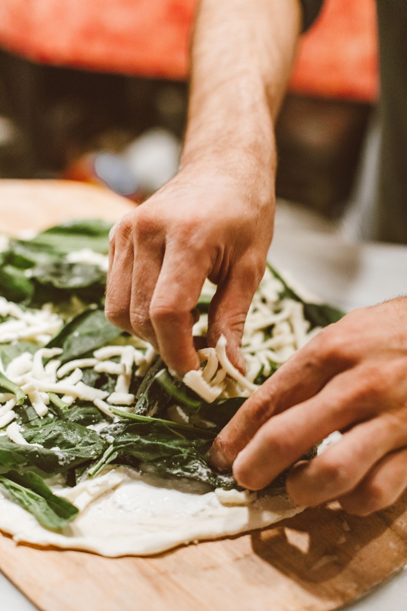 Detailed close-up of hands placing fresh spinach and cheese toppings on pizza dough for cooking.