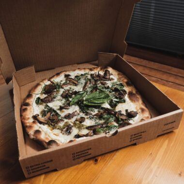 Freshly baked thin crust pizza with mushrooms and mozzarella in a cardboard box on a wooden table.